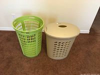 Two plastic clothes baskets side by side on carpet, one green with open mesh and the other beige with lid.