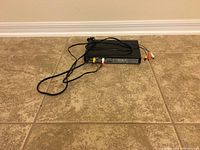 Front and top view of black Sony DVD player placed on tiled floor with power cord and AV cables visible.