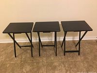 Three small black folding tables standing upright on tiled floor against a light wall