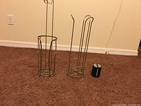 Two metal holders for toilet paper rolls and one metal toothbrush holder sitting on carpet floor near wall outlet.