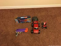 Photo showing the red remote-controlled truck, the projectile toy in box, the small ATV toy, and the Fortnite projectile launcher outside of box on carpet.