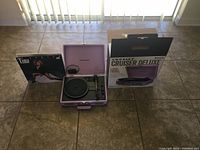 Portable lavender Crosley Cruiser Deluxe turntable displayed with a Tina Turner vinyl album and original product box behind, showing all included items and packaging.