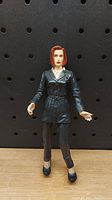 Photo of the Agent Scully figure standing against a black pegboard background, clear front view showing details of clothing and facial features.
