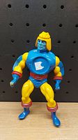 Front view of vintage Masters of the Universe figure showing yellow skin, blue armor, red accents, and a worn sticker on chest.