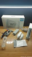 Photo of Nintendo Wii console with Wii Remote, Nunchuk, power adapter, AV cables, clear console stand, instructions, and original Wii box in background.