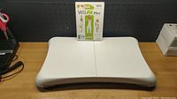 Photo of Nintendo Wii Balance Board on a wooden surface with Wii Fit Plus game case propped up behind it.