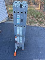 Folded ladder standing on asphalt showing its hinged joints and orange safety foot pads.
