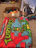 Wide shot showing the Teenage Mutant Ninja Turtles sleeping bag spread out with a box full of assorted action figures behind it, part of another basket of toys also visible.