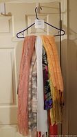 Scarfs hung on a hanger showing different colors and textures including peach lace, white, orange fringed, and multicolored floral patterns.