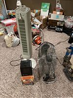 Photo of standing tower fan, ceramic heater in box, and high velocity fan in a cluttered basement room.