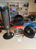 Three black and grey electric heaters and fans: Lasko tower heater, Honeywell Turbo Force fan with box, and black Vornado fan on tabletop.