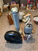 Full view of the three appliances on floor including black Honeywell fan, white Windcrest tower heater, and Lasko heater on box.