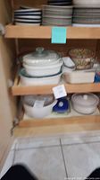 Two shelves of assorted bowls and platters inside a cabinet, showing Corningware dishes and ceramic bowls, some stacked and needing cleaning.