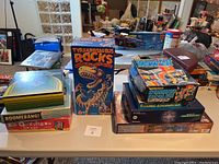 Photo showing full assortment of 8 games stacked on a table, featuring Tyrannosaurus Rocks, Domino Dealer, Cosmic Encounter, Space Chess, Boomerang, and 20 Questions boxes.