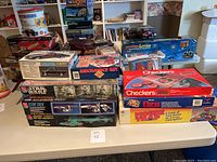 Wide view of table with multiple boxed board games and model kits stacked, showing Checkers, Chess, Mouse Trap game boxes, Star Wars and Star Trek model kits, and a Chevrolet truck model kit.
