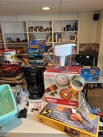 Photo shows multiple items grouped on table including a black Cuisinart drip coffee maker, Nesco food dehydrator box, Brita water pitcher, Oster foot massager box, and silver desk lamp.