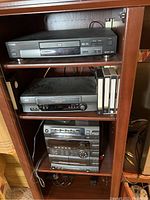 Shelf with Toshiba DVD player on top, JVC VHS player with four VHS tapes in middle, and Sony stereo system on bottom shelf.