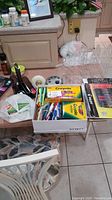 Box with assorted office items on kitchen counter including Crayola crayons, Sharpie markers, pens, pencils, noise machine