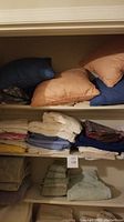 Photo showing top shelf with pillows in navy blue and peach colors, middle and bottom shelves with folded towels and linens in multiple colors including white, blue, yellow, and beige.