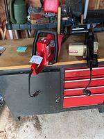 Sears red electric hedge trimmer and Craftsman electric chainsaw on wooden and metal workbench in shed. Both have power cords and visible cutting blades.