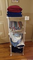 Full view of white metal 3-tier storage rack with assorted folded linens and blankets on each shelf and on top.