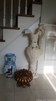 Three decorative items arranged side-by-side against staircase wall: large cream pitcher statue, blue two-handled vase, gold bowl with lid.