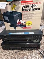 Photo showing Ambico Deluxe Video Transfer System box with JVC and Memorex VHS players stacked on the floor.