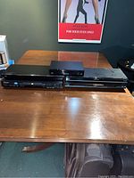 Four DVD players on a wooden table. Three front views show the DVD players stacked horizontally with visible disc trays and front panels.