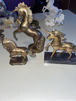Photo of brass unicorn statues on different bases and white ceramic unicorn horse figurines in background.