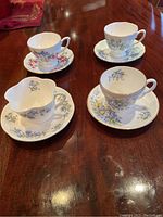 Four tea cup and saucer sets arranged on a wooden table, showing floral patterns in blue and pink, different cup rim styles.