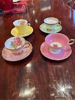 Lot of 4 tea cups and saucers on wooden table showing different colors and floral designs of each cup and saucer set.