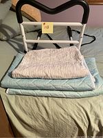 Front view of adjustable bed assist rail with black padded M-shaped grip and attached straps, placed on a bed with protective pads underneath.