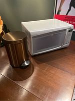 White Panasonic microwave and rose gold stainless steel step trash can on a wooden surface.