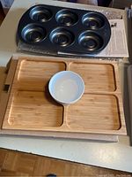 Black metal brookie pan with six rounded muffin cups and rectangular bamboo serving tray divided into four sections with a small white ceramic bowl in the center.
