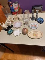 Photo showing assorted decorative items including white hands figurines on black bases, mugs with animal motifs, floral decorated plate, pink and white ruffled dishes, marble-effect candle holders, and blue glass orb