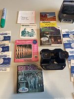 Photo showing Sawyer View-Master viewer, multiple View-Master reels and original packets with various themes such as Lost in Space, Green Hornet, and Rockefeller Center.