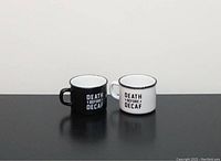 Two cappuccino mugs side by side, one black and one white with 'Death Before Decaf' text, shown on a flat dark surface with plain background.