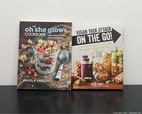 Photo of two vegan cookbooks: "The Oh She Glows Cookbook" and "Vegan Yack Attack On the Go!" standing upright side by side on a black surface against a plain background.