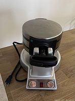 Side view of the closed Cuisinart Waffle and Omelette Maker with power cord visible, showing the control panel with two dials and a power switch.