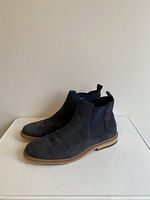 Side and angled view of a pair of men's blue suede desert boots with elastic side panels and light brown soles on white surface against a plain wall.