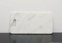 Photo of rectangular white marble cutting board with rounded corner and oblong hole for hanging. Marble has natural subtle veining. Felt pads visible on one side to prevent slipping.