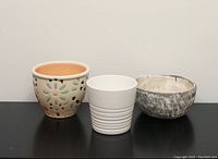 Front view of three decorative small plant pots: multicoloured floral, plain white ribbed, and grey rustic finish.