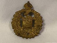 Close-up photo of brass cap badge showing overall shape, crown at the top, circular frame, and central shield with crossed rifles and lion.