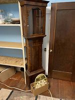 Full view of the wooden grandfather clock case with glass door, showing the cardboard box with the clock dial face and parts at the base.