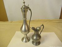 Full view of two vintage pewter serving jugs on a beige cloth background, showing size and shape difference.