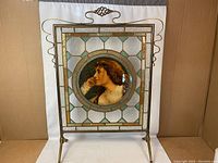 Full view of the fire screen, showing the metal frame, stand, and stained glass panel with circular portrait in the center.
