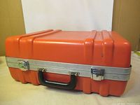 Closed view of the bright orange hard plastic carry case with metal trim and black handle.