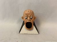 Front view of the ceramic figurine with a human head and multiple small holes on top; mounted on a black and white triangular base. Mouth open areawhere toothpicks can be placed.