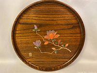 Full view of round wooden plate with painted floral design of orange and purple flowers and green leaves on wood grain background.