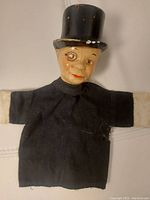 Front full view of the black fabric hand puppet body with beige wooden head wearing a black top hat.